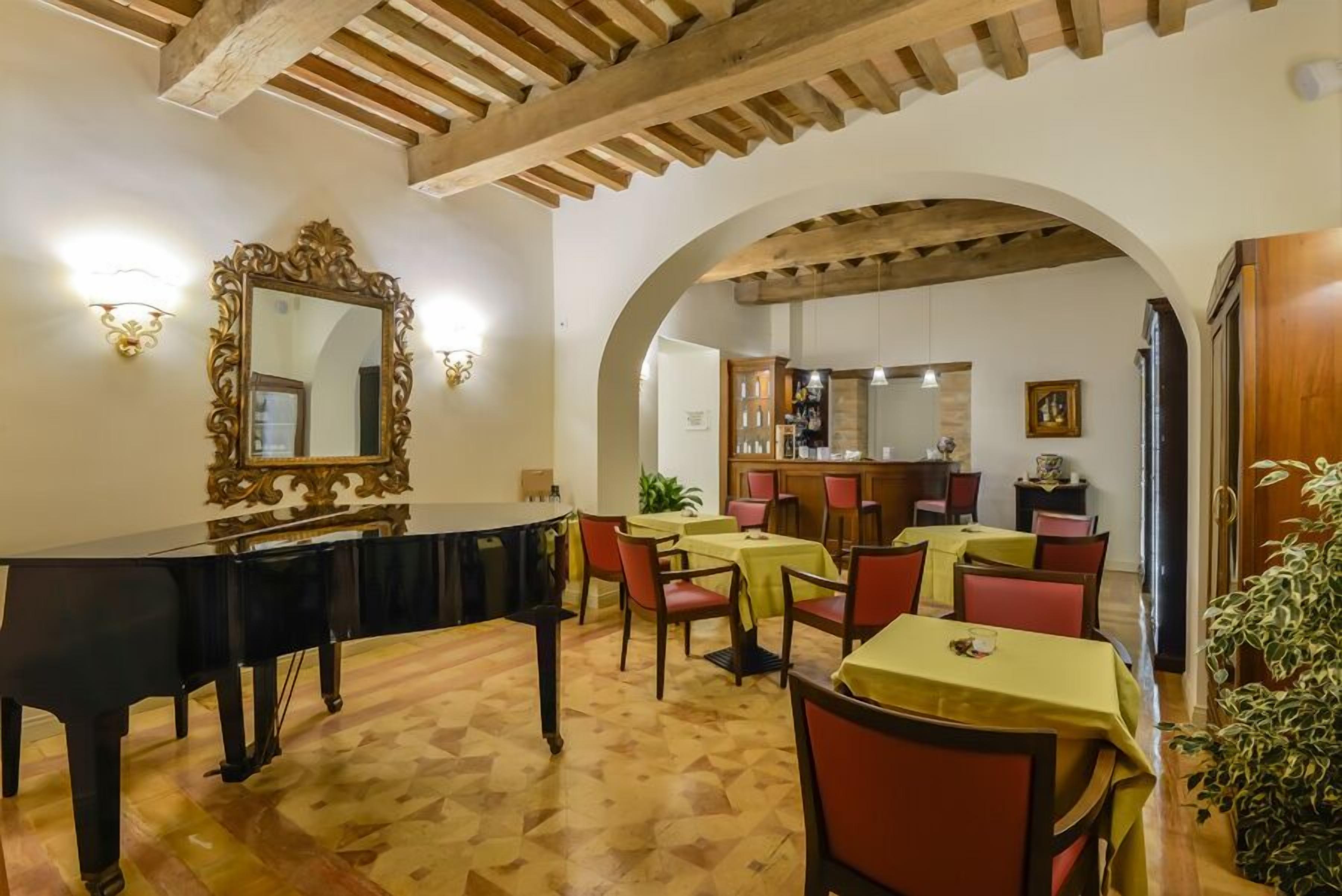 Un comedor con un piano de cola, vigas de madera y un espejo grande. Las mesas están dispuestas con sillas, y se observa una planta.