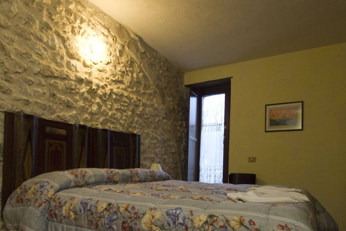 A bedroom with a stone wall, a bed with floral bedding, a window with curtains, and a framed picture on the wall.