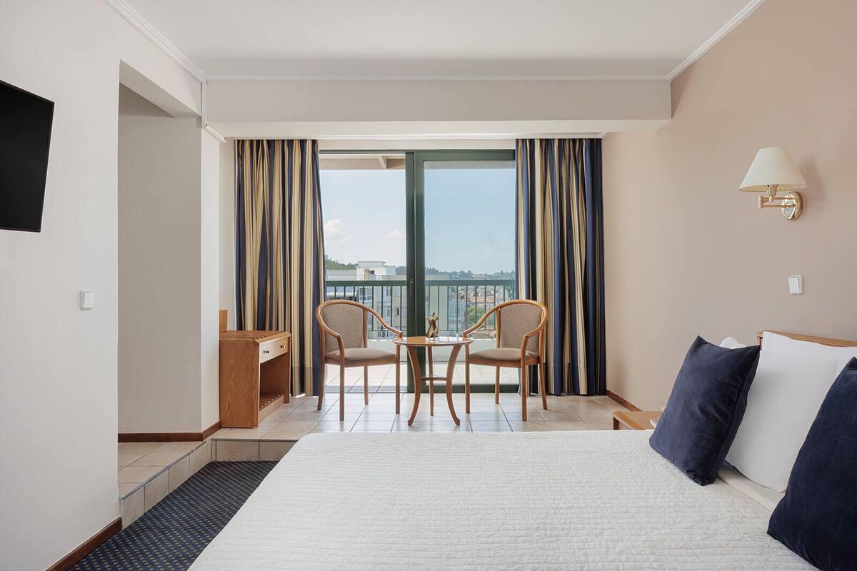 A hotel room with a bed, two chairs, a small table, a desk, a TV, and a balcony with curtains.