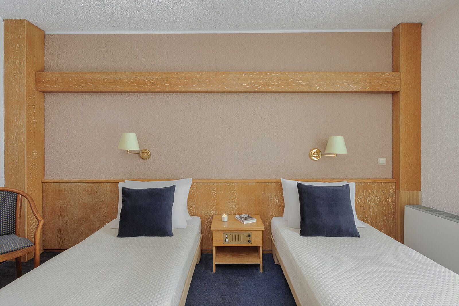Quarto de hotel com duas camas de solteiro, cabeceira de madeira, mesa de cabeceira com luminária e uma cadeira.