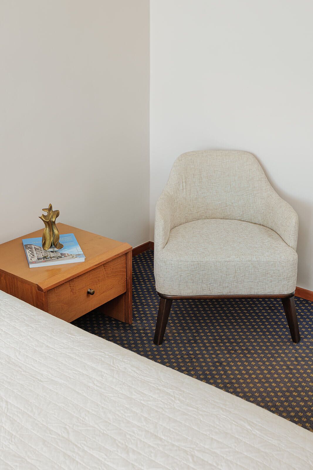A hotel room with a bed, a small wooden nightstand with a decorative item, a light-colored armchair, and a patterned carpet.