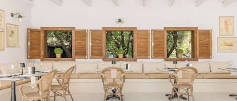 A dining area with a long table, wicker chairs, and a bench with cushions. There are windows with wooden shutters and potted plants.