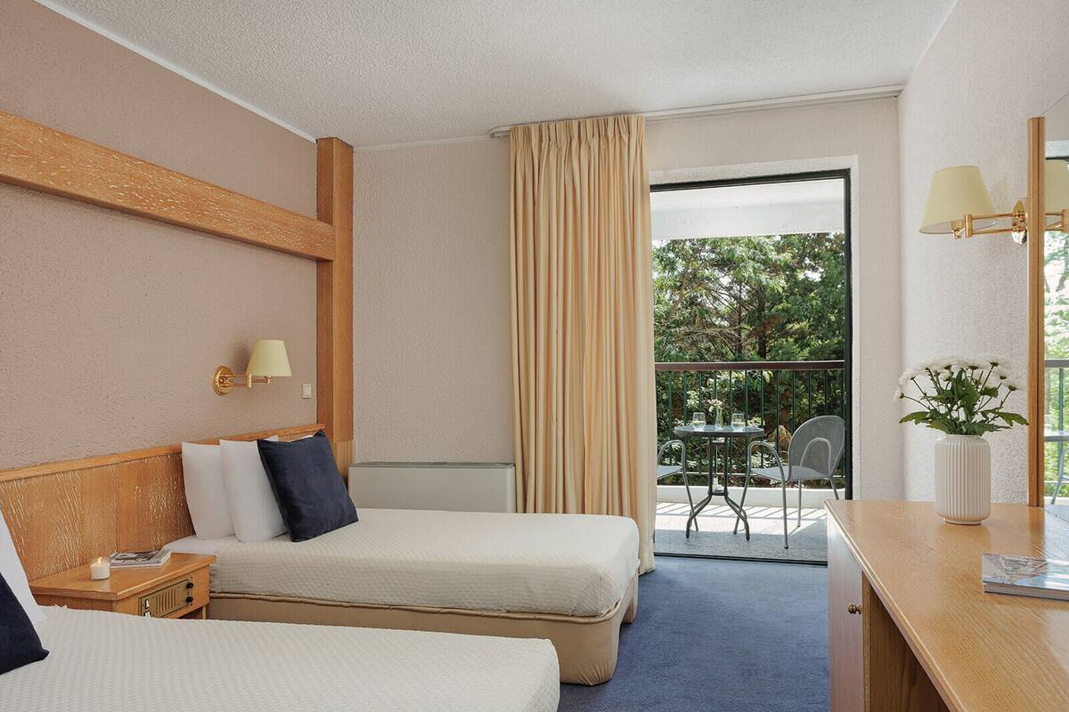 A hotel room with two beds, a wooden headboard, a nightstand, a lamp, a window with curtains, and a balcony with a table and chairs.