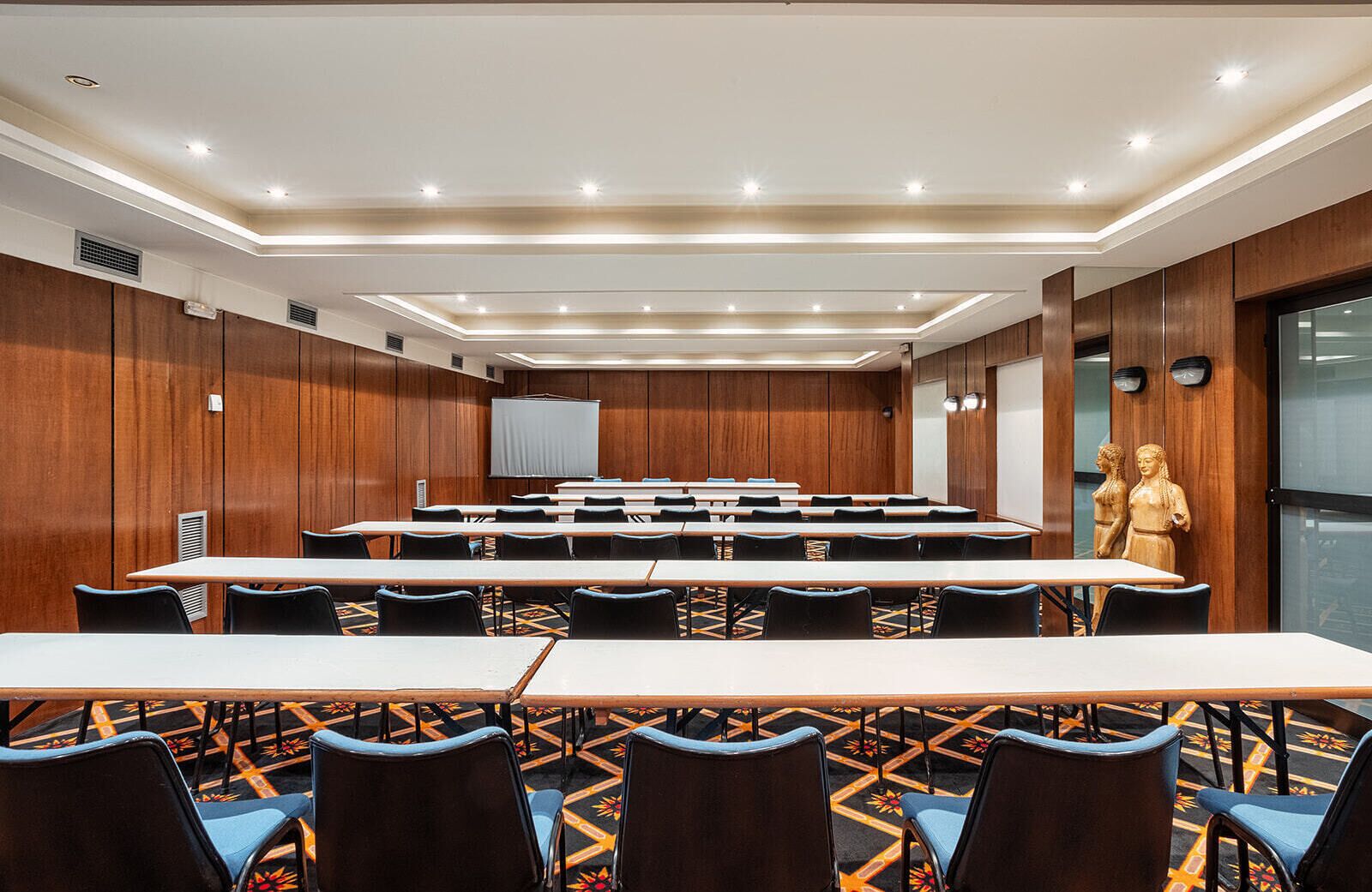 Un salón de conferencias con largas mesas y sillas, paneles de madera y una gran pantalla al frente.