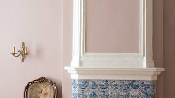 A room with a pink wall, a white mantelpiece above a blue and white tiled fireplace, and a wooden chair with floral upholstery.