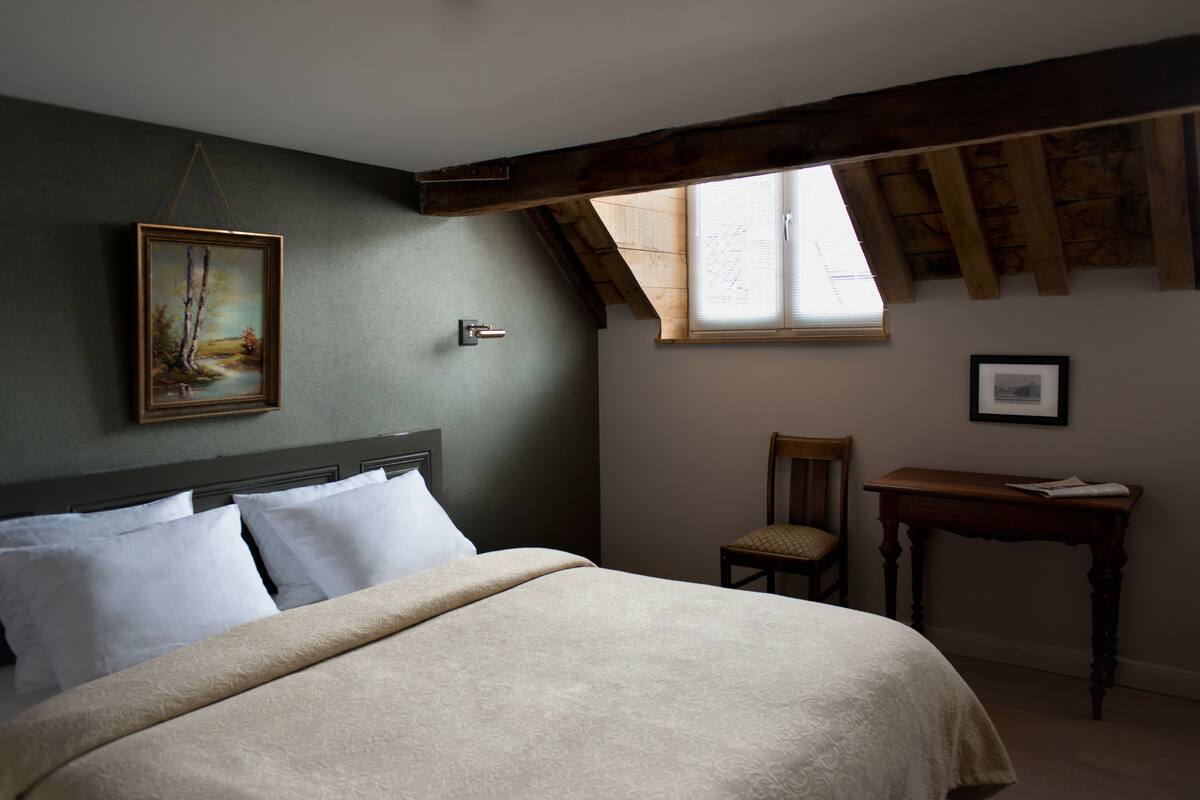 A bedroom with a bed, a chair, a table, a painting, and a window.