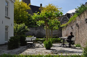 Une cour avec un bâtiment blanc, un mur de briques et un arbre. On y trouve des bancs et une table avec des chaises.