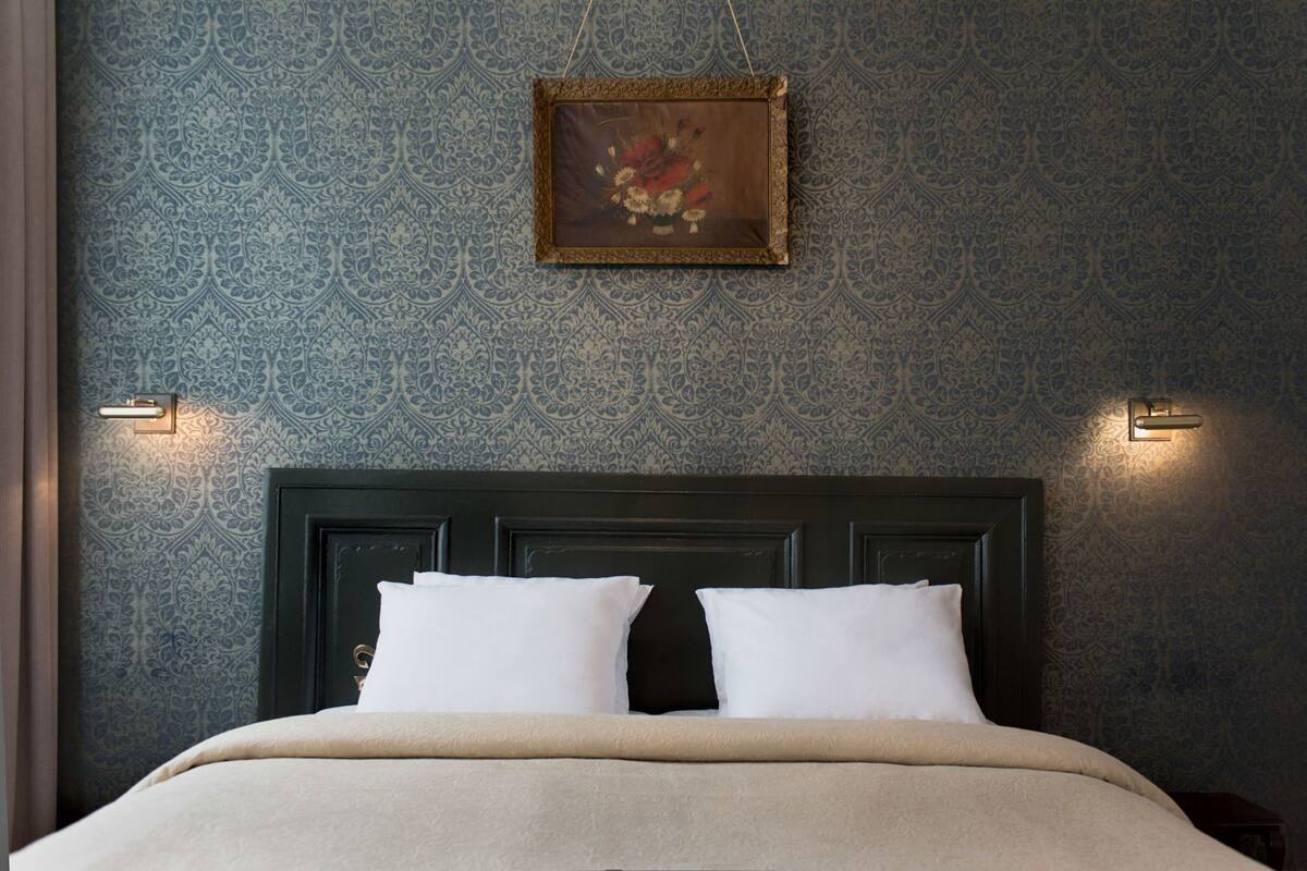 A bed with white pillows and a dark headboard, a framed picture above, and wall sconces on either side.