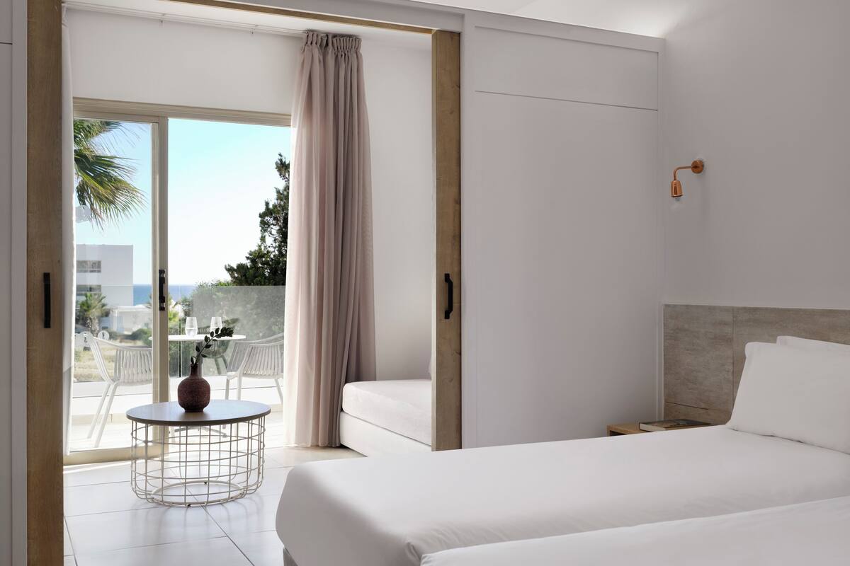 A modern hotel room with a large bed, a round table with a vase, and a balcony with a view of the sea.