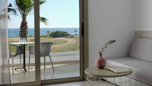 Une chambre moderne dotée d’une porte-fenêtre coulissante donnant sur un balcon avec vue sur la mer et un palmier. On y trouve une table ronde avec un vase et un livre ouvert, ainsi qu’un canapé avec un coussin.