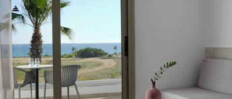A modern room with a sliding glass door leading to a balcony with a view of the sea and a palm tree. There is a round table with a vase and an open book, and a sofa with a cushion.