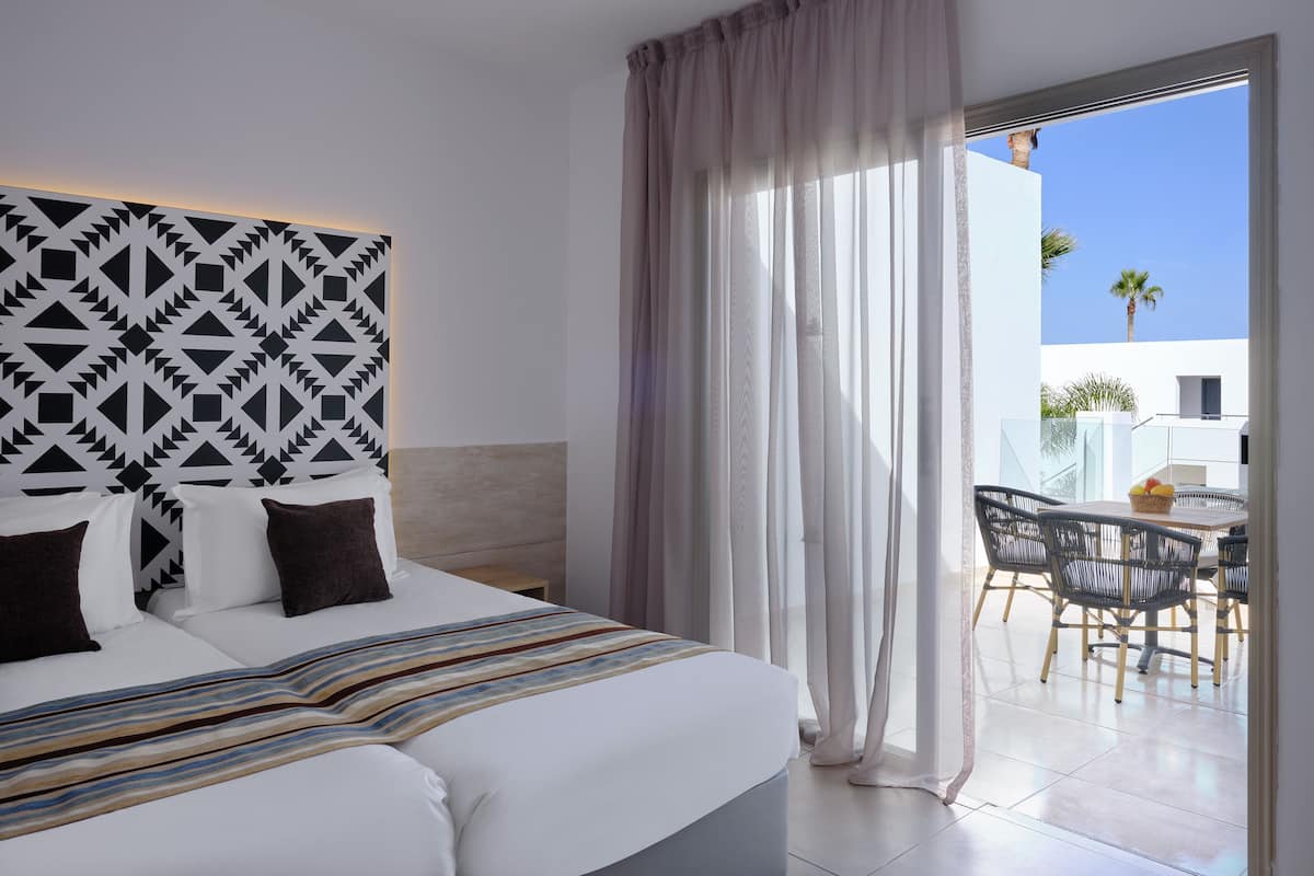 A modern hotel room with a large bed, a geometric-patterned headboard, a balcony with a table and chairs, and a view of palm trees.