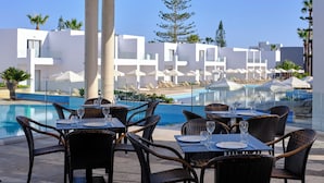 Un espace de restauration aménagé au bord de la piscine, avec des tables et des chaises, une piscine, et des bâtiments modernes en arrière-plan.