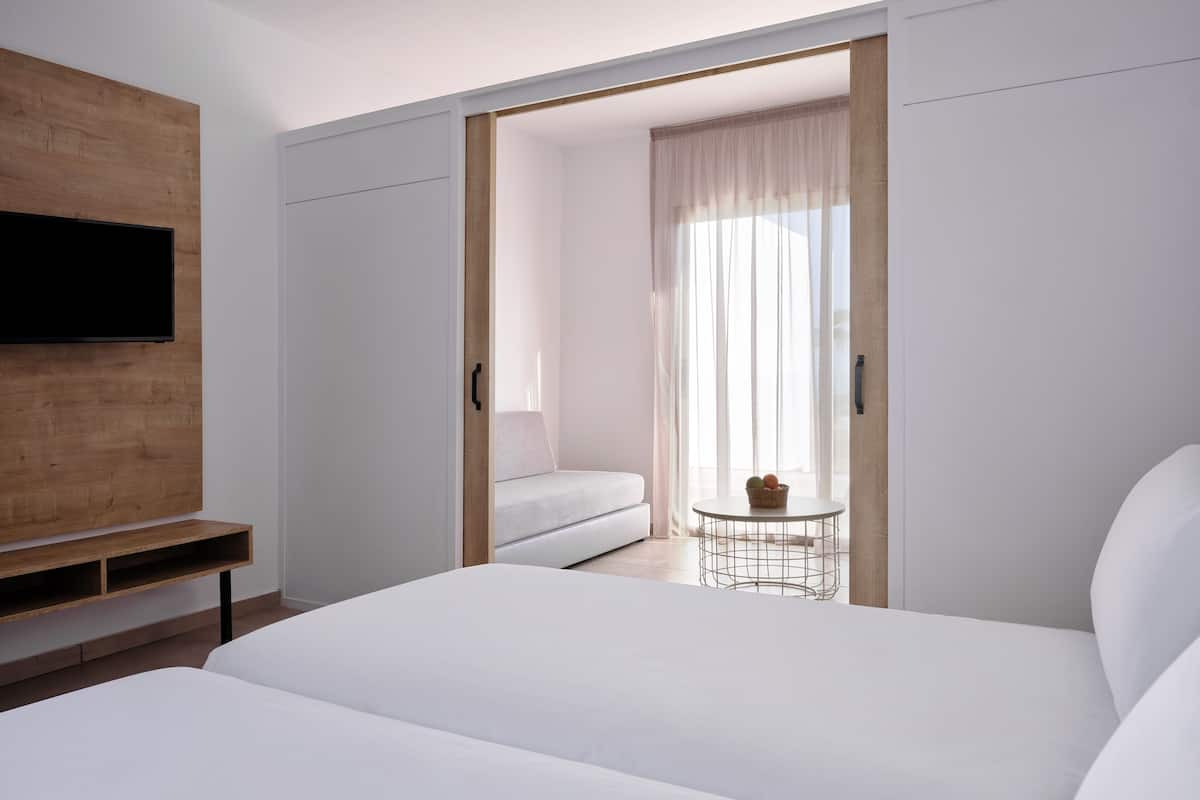 A modern hotel room with a large bed, a TV mounted on a wooden panel, a sofa, and a small table with a basket.