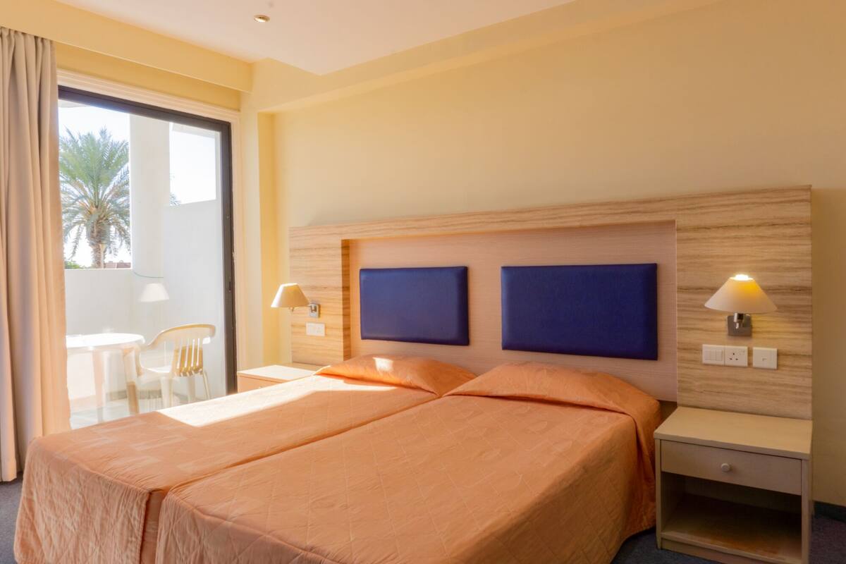 A hotel room with a bed, two bedside lamps, a nightstand, and a view of a balcony with a chair and palm tree.