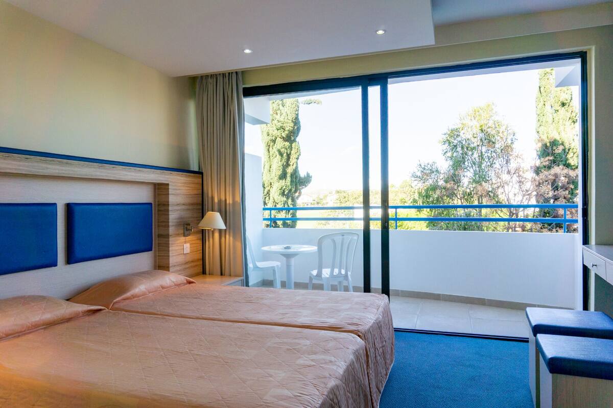 A hotel room with two beds, a desk, a chair, and a balcony with a view of trees and a blue railing.