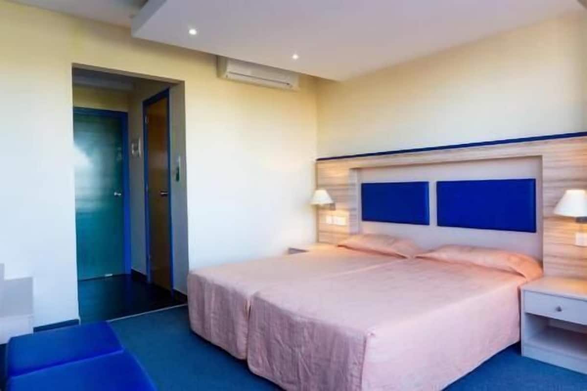 A hotel room with a bed, bedside tables, a nightstand, a door, and an air conditioning unit.