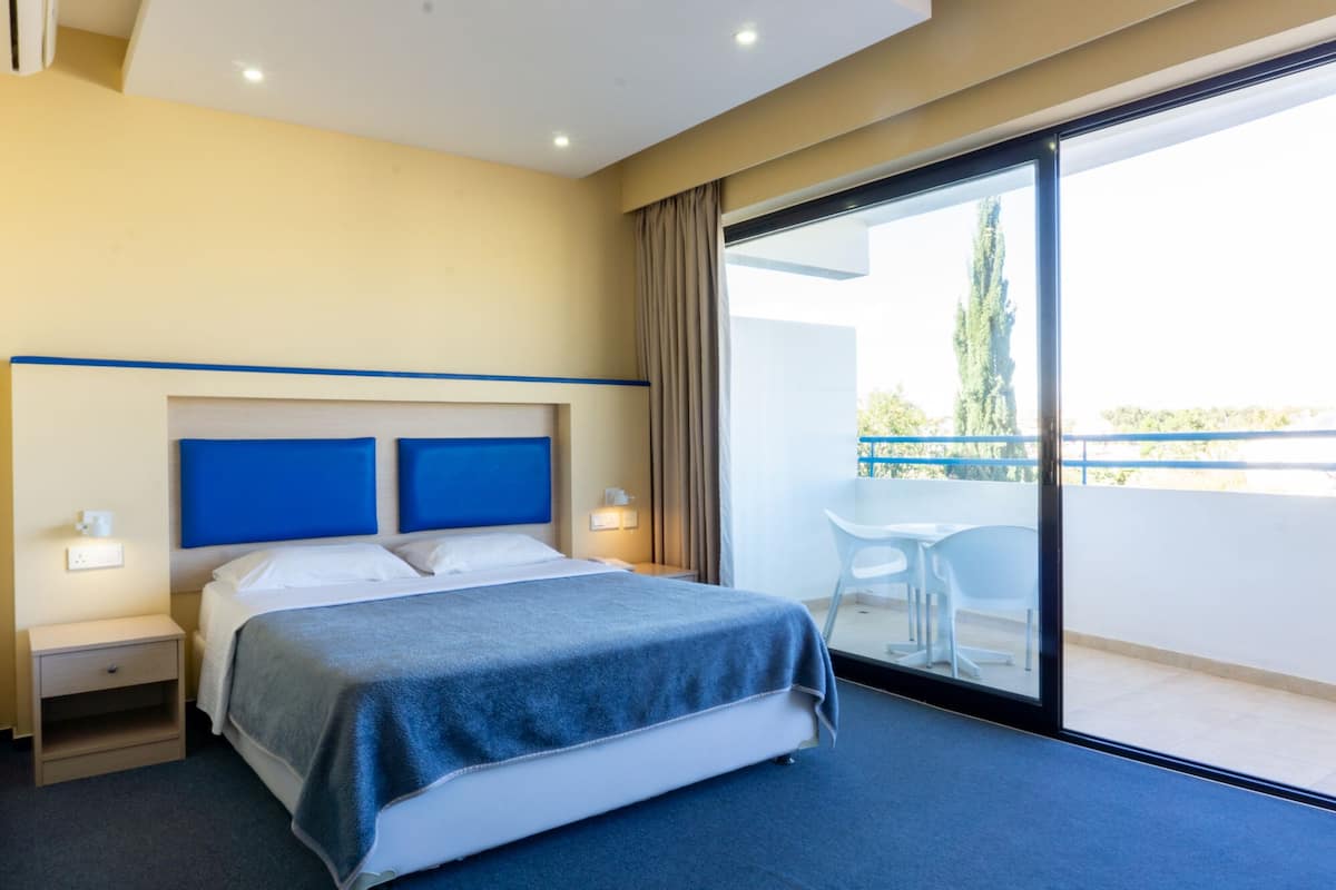 A hotel room with a bed, bedside tables, a nightstand, a window with a view of the outdoors, and a balcony with a table and chairs.
