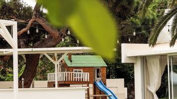 Children can play on the outdoor play structure surrounded by lush greenery and a building with a green roof.
