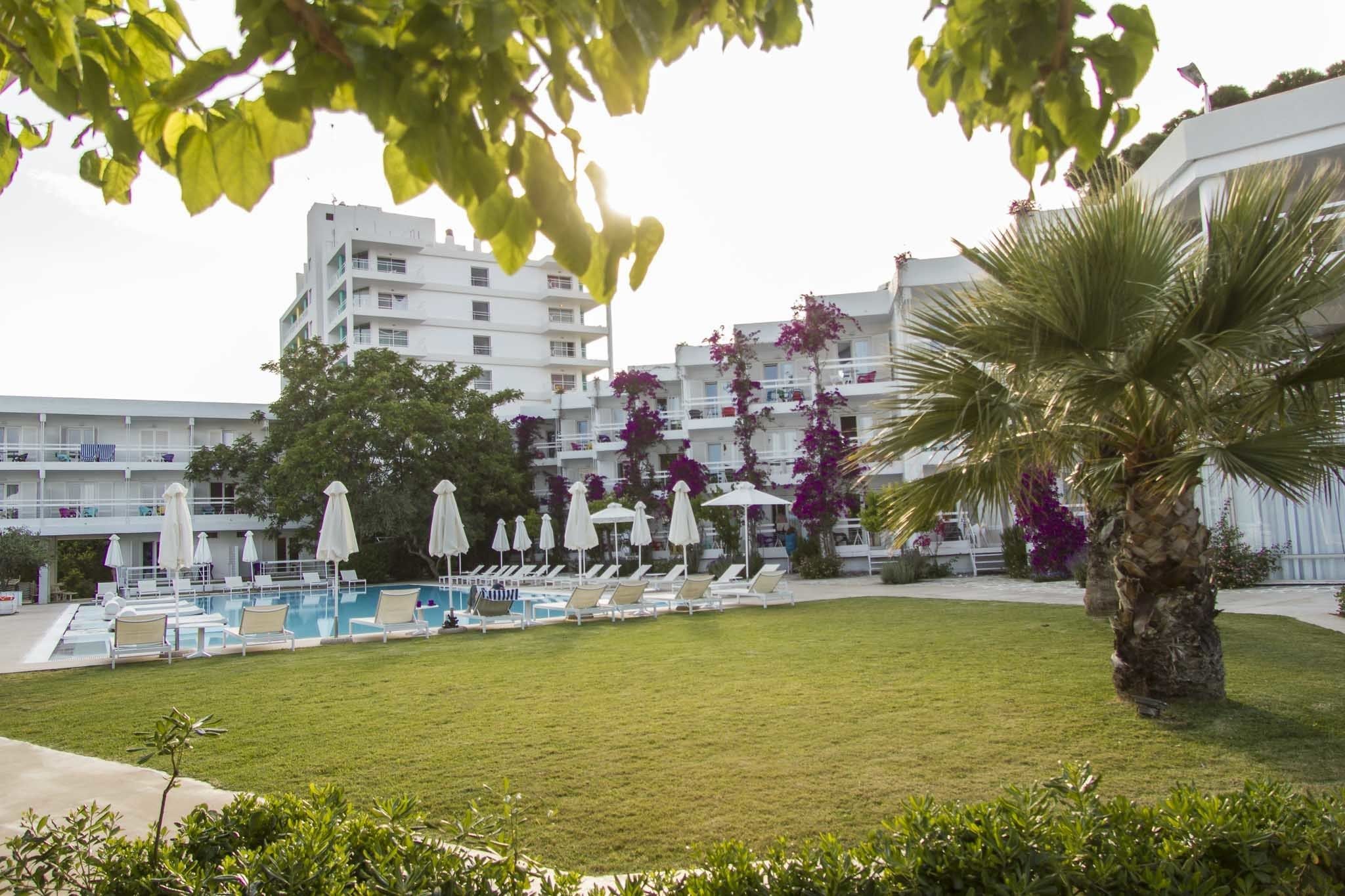 Un área de piscina con sillones de descanso y sombrillas, rodeada de vegetación y un edificio con balcones.