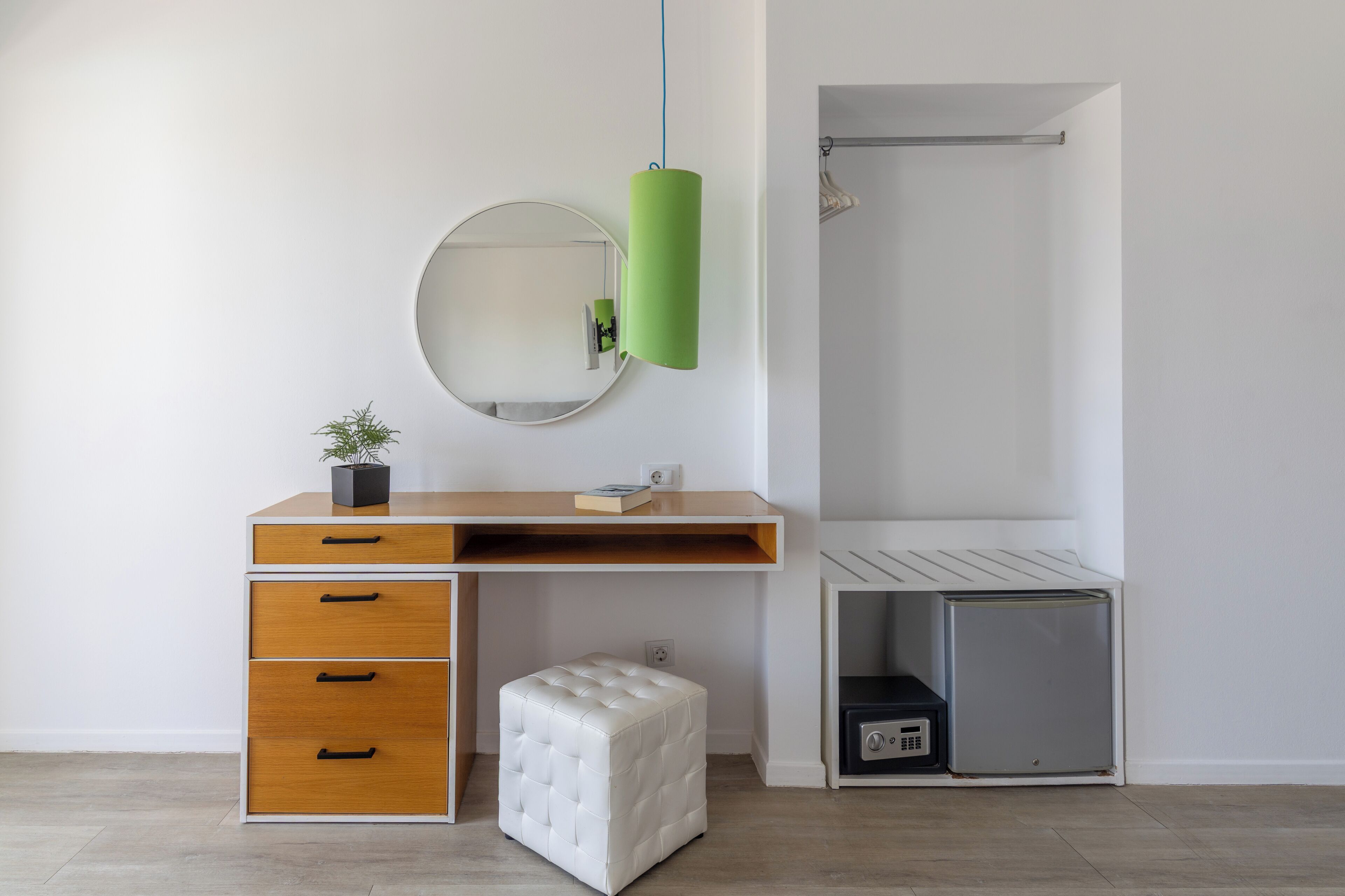 Un dormitorio moderno con un escritorio de madera, un espejo redondo, una lámpara colgante verde, un reposapiés blanco y un vestidor.