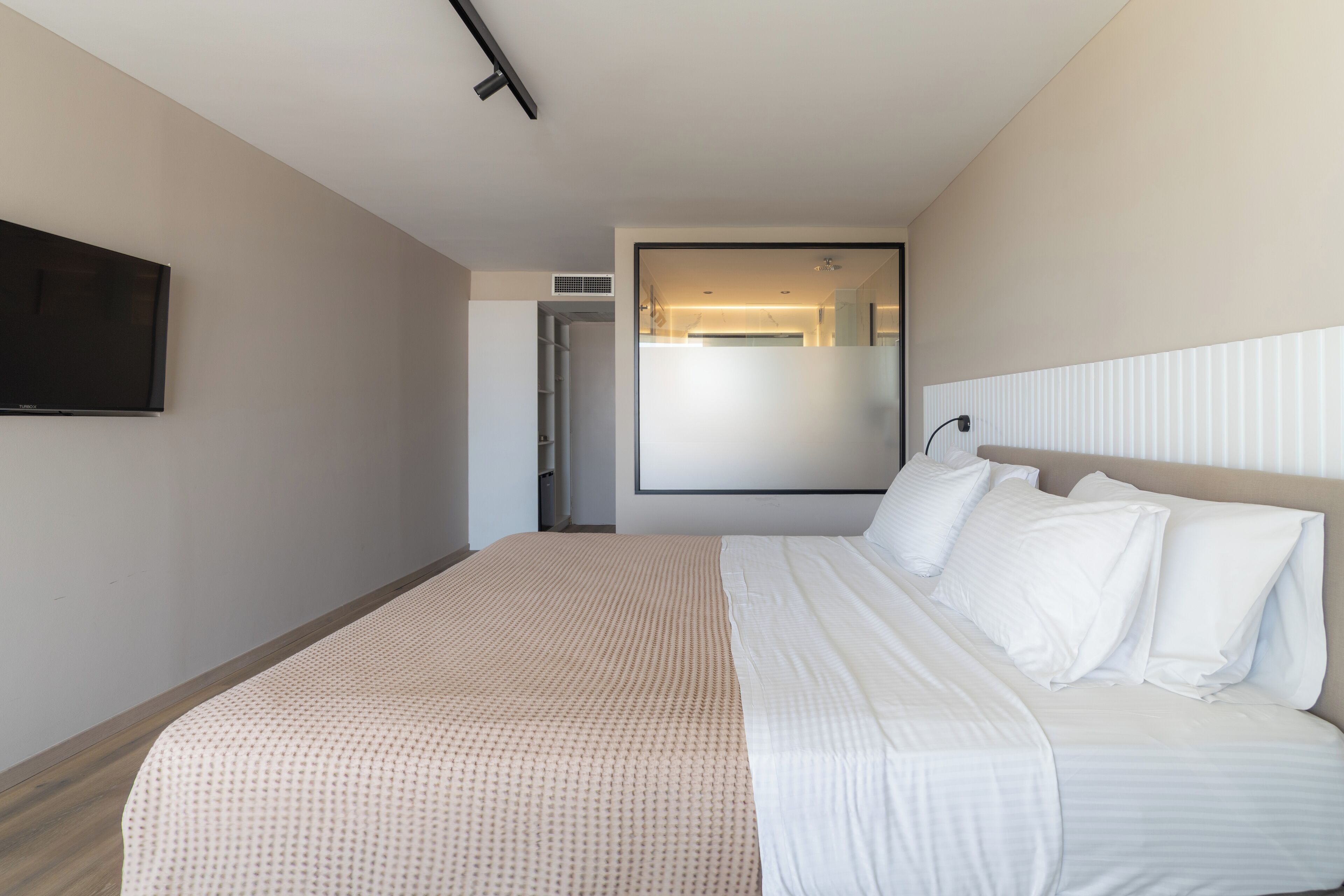 A modern hotel room with a large bed, a flat-screen TV mounted on the wall, and a large window providing natural light.