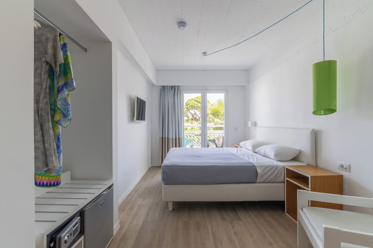 A bedroom with a bed, a small bedside table, a hanging green light fixture, a window with curtains, and a view of the outdoors.