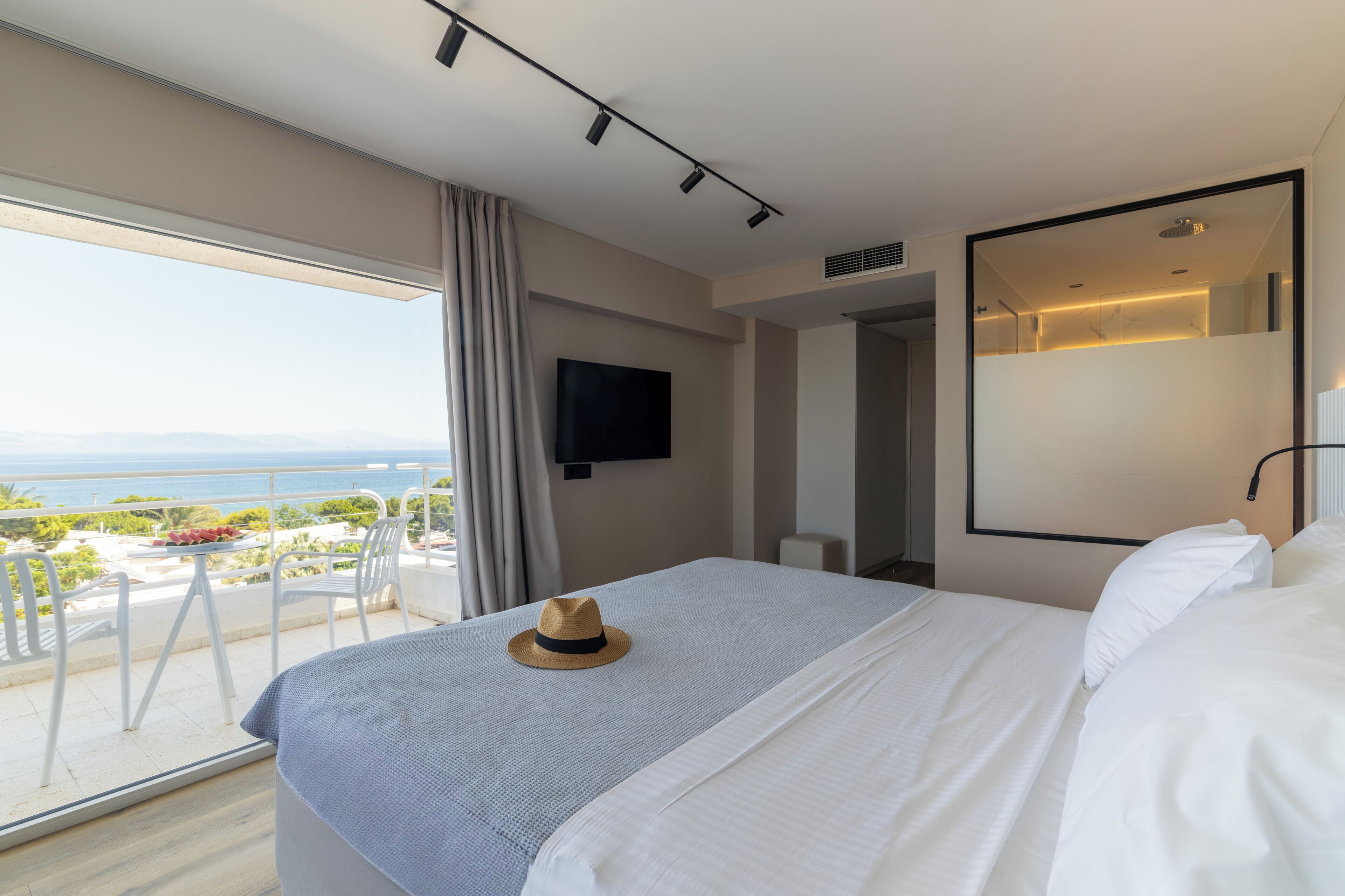 A hotel room with a bed, a TV, a balcony with a view of the sea, and a dining table with a hat on it.
