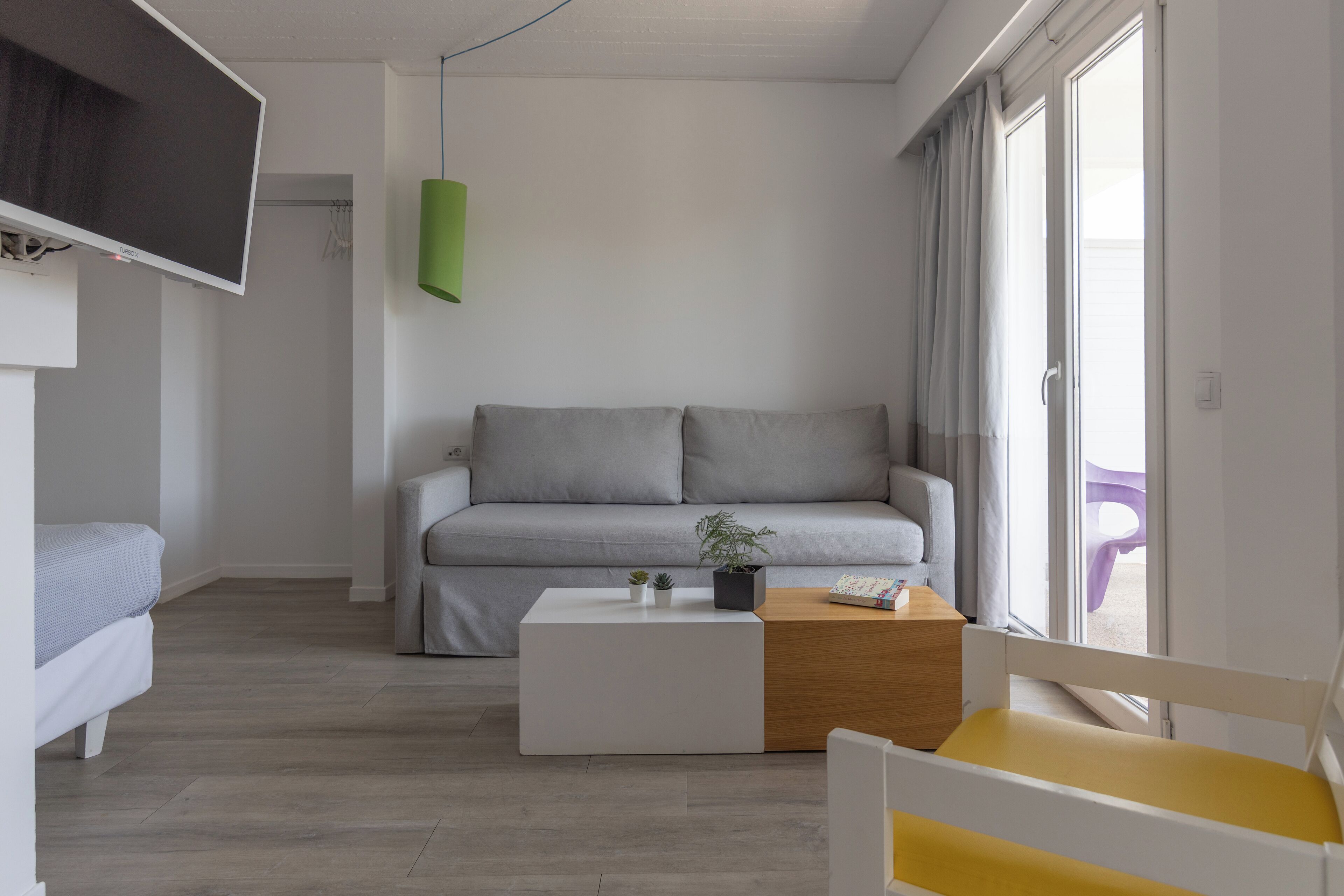 Una sala de estar moderna con un sofá gris, una mesa de centro blanca, una mesita de noche de madera, un televisor montado en la pared y una ventana con cortinas.