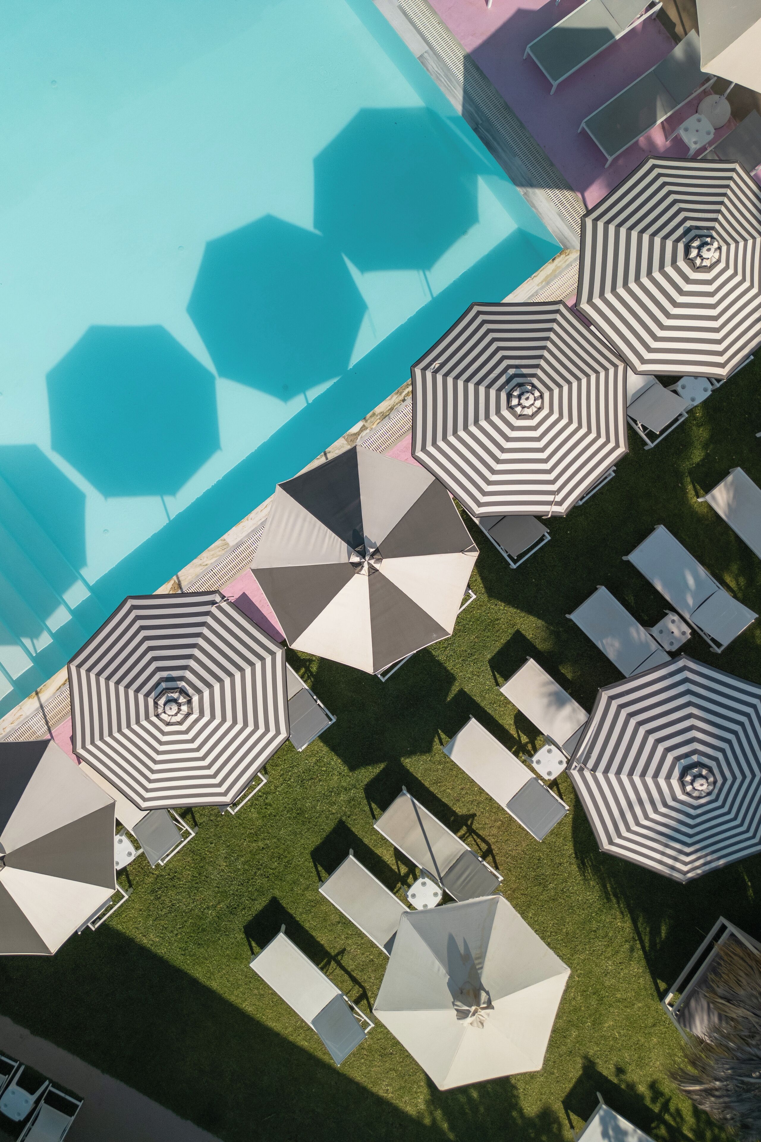 Vue aérienne d’un espace autour d’une piscine, avec des carreaux hexagonaux recouvrant le fond de la piscine, plusieurs chaises longues et des parasols à rayures.