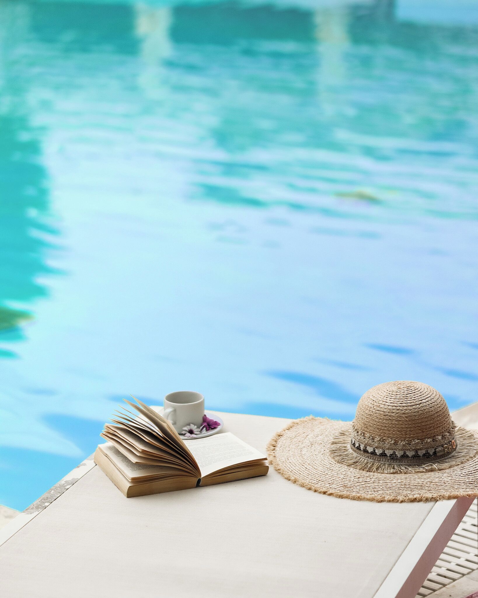 A poolside setting with a book, a cup, and a straw hat on a white surface.