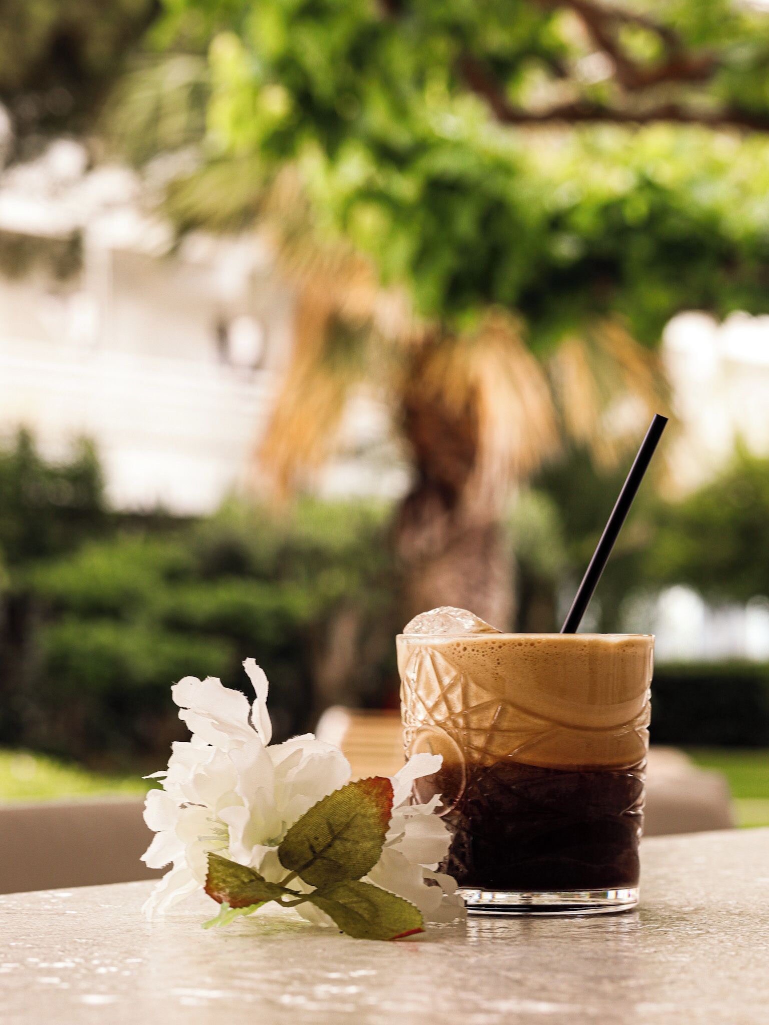 A glass of iced coffee with a straw on a table, accompanied by a white flower.