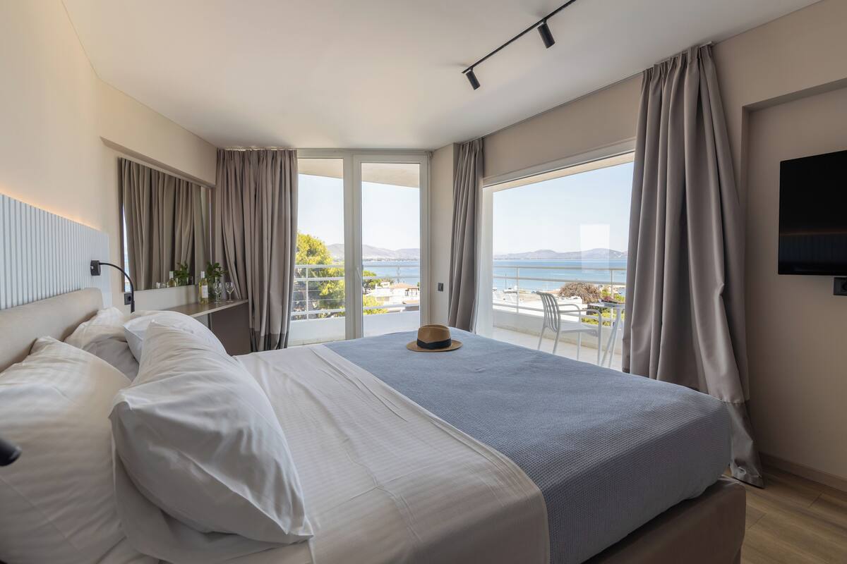 A modern hotel room with a large bed, a TV, and a balcony with a view of the sea.