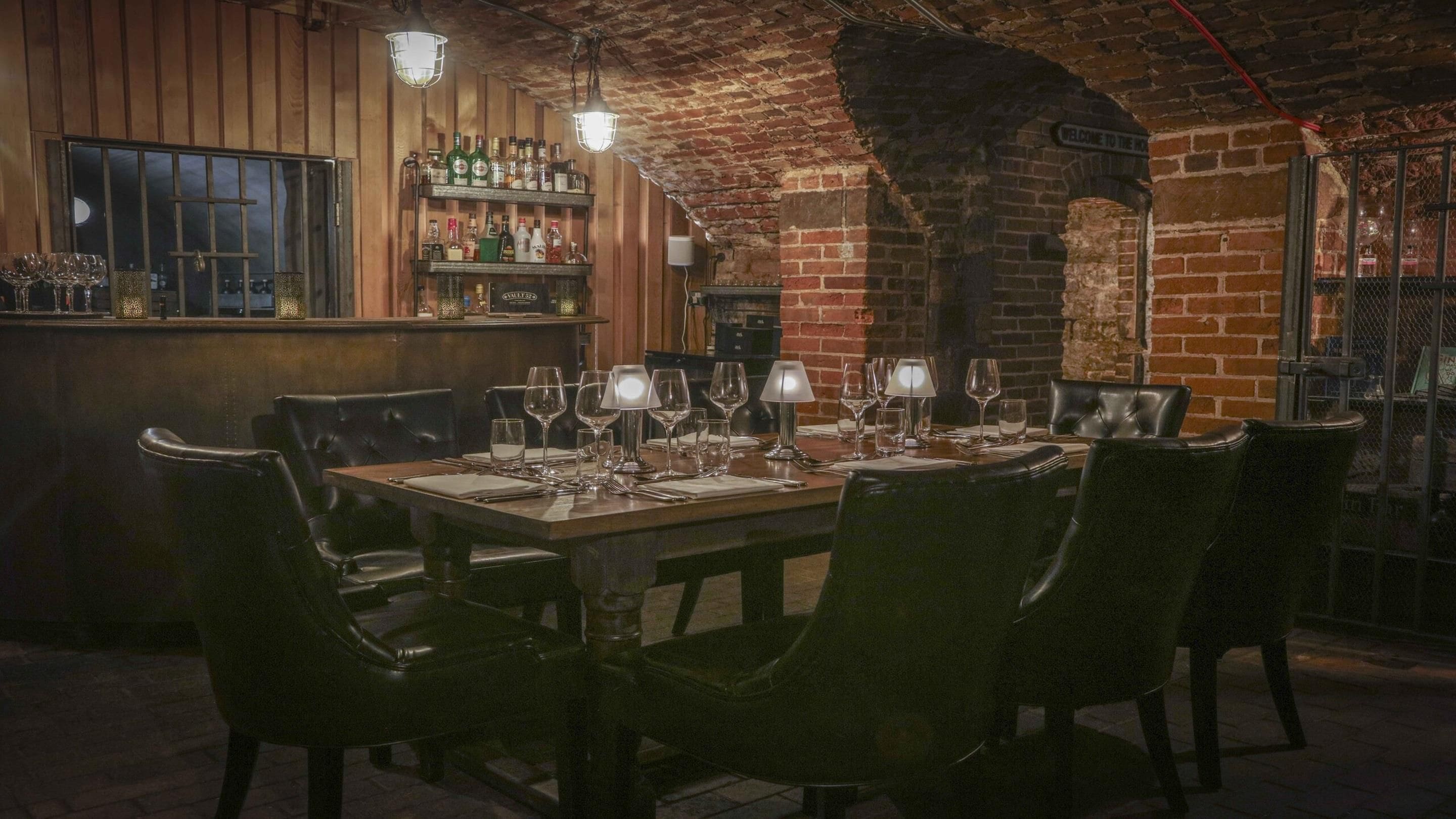 A brick-walled bar area with a wooden counter and shelves of bottles.
