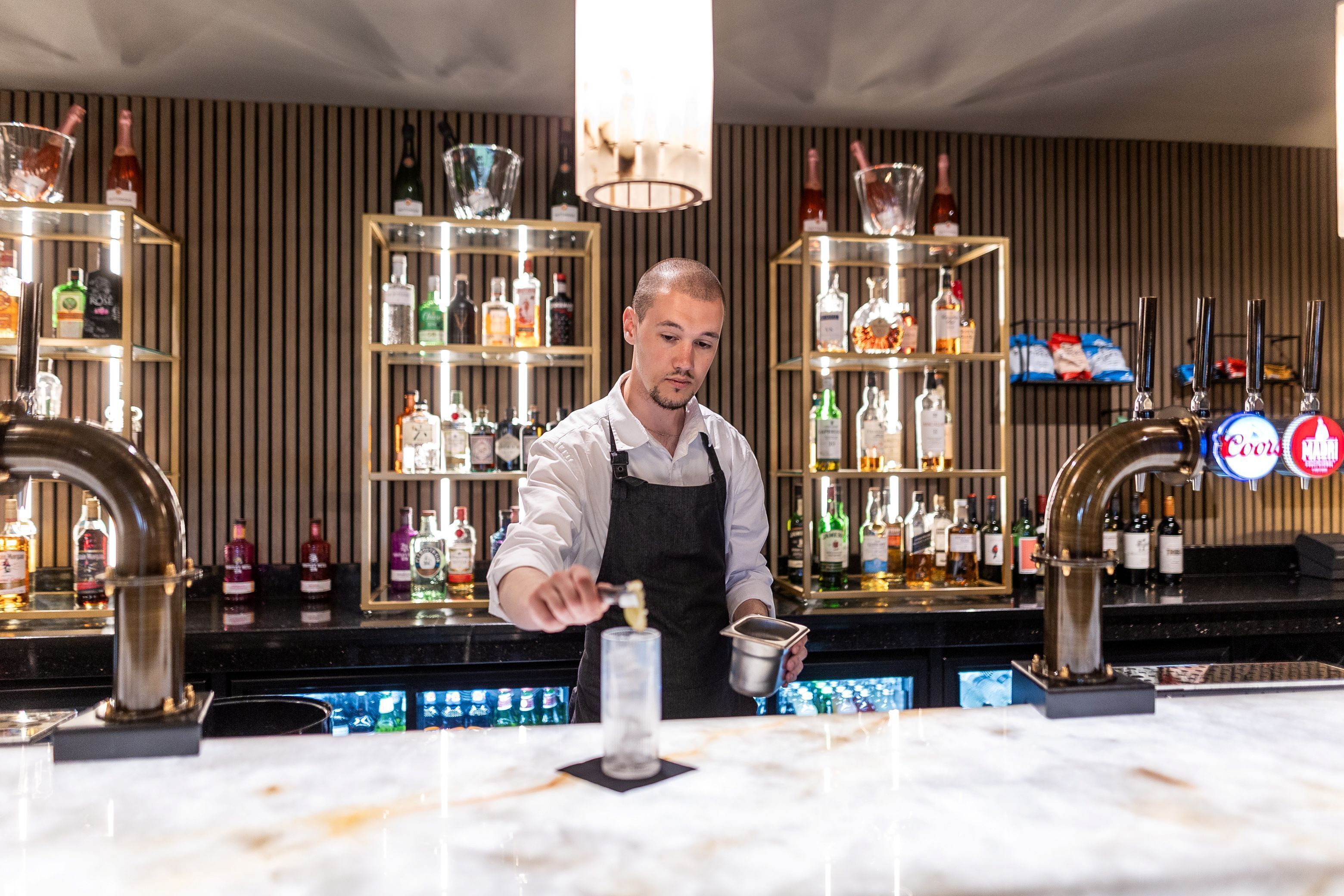 A bar area with shelves of liquor bottles, a bartender preparing a drink, and a bar counter with taps and a cash register.