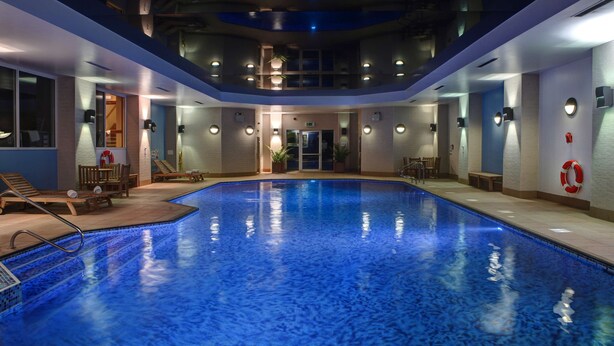 An indoor swimming pool with a blue mosaic tile floor, surrounded by lounge chairs and tables, and equipped with ceiling lights and wall-mounted lamps. -  Windmill Village Hotel, Golf Club & Spa, BW Signature Collection (Coventry)