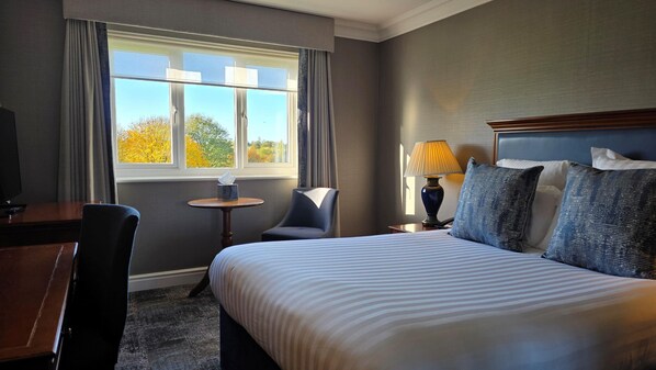 A hotel room with a large bed, a desk, a chair, a small table, and a lamp.