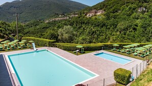 Un área de piscina con dos piscinas, rodeada de sillas y sombrillas verdes, con un telón de fondo de colinas verdes y edificios.