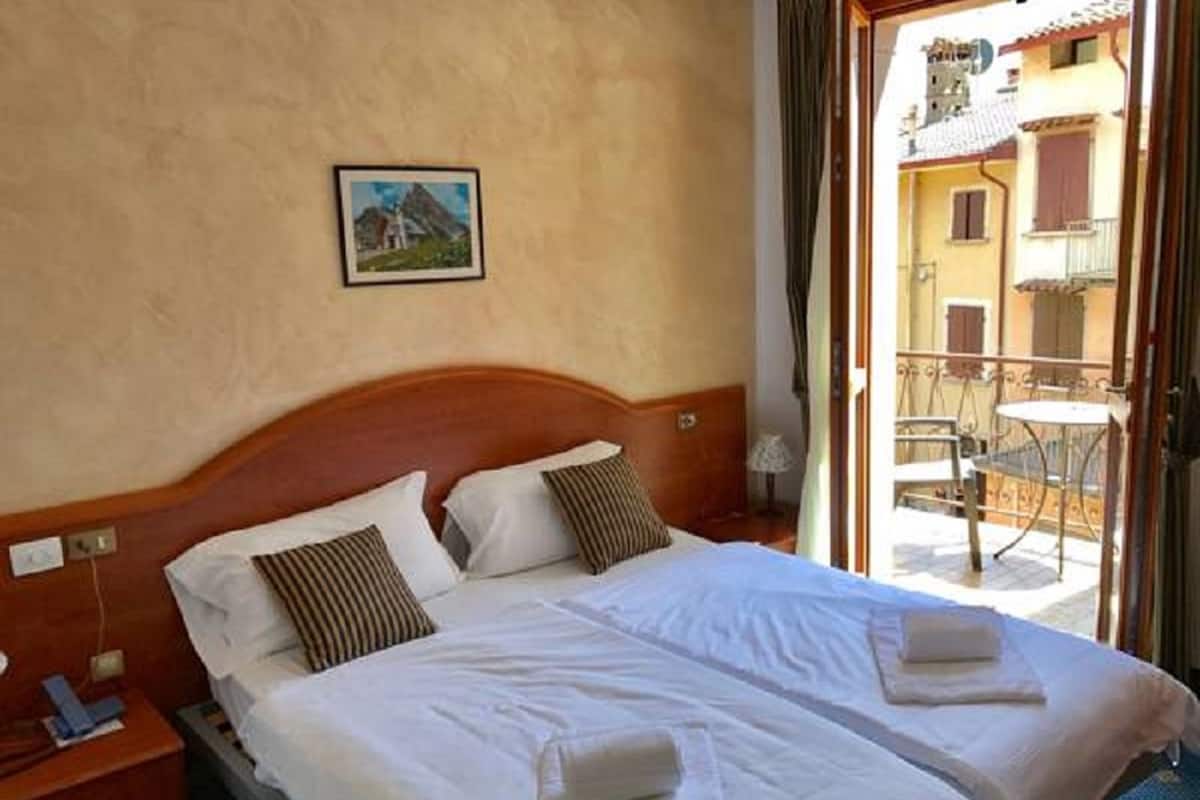 A double bed with white linens and two pillows, a wooden headboard, a bedside table with a lamp, and a view of a balcony with outdoor furniture.