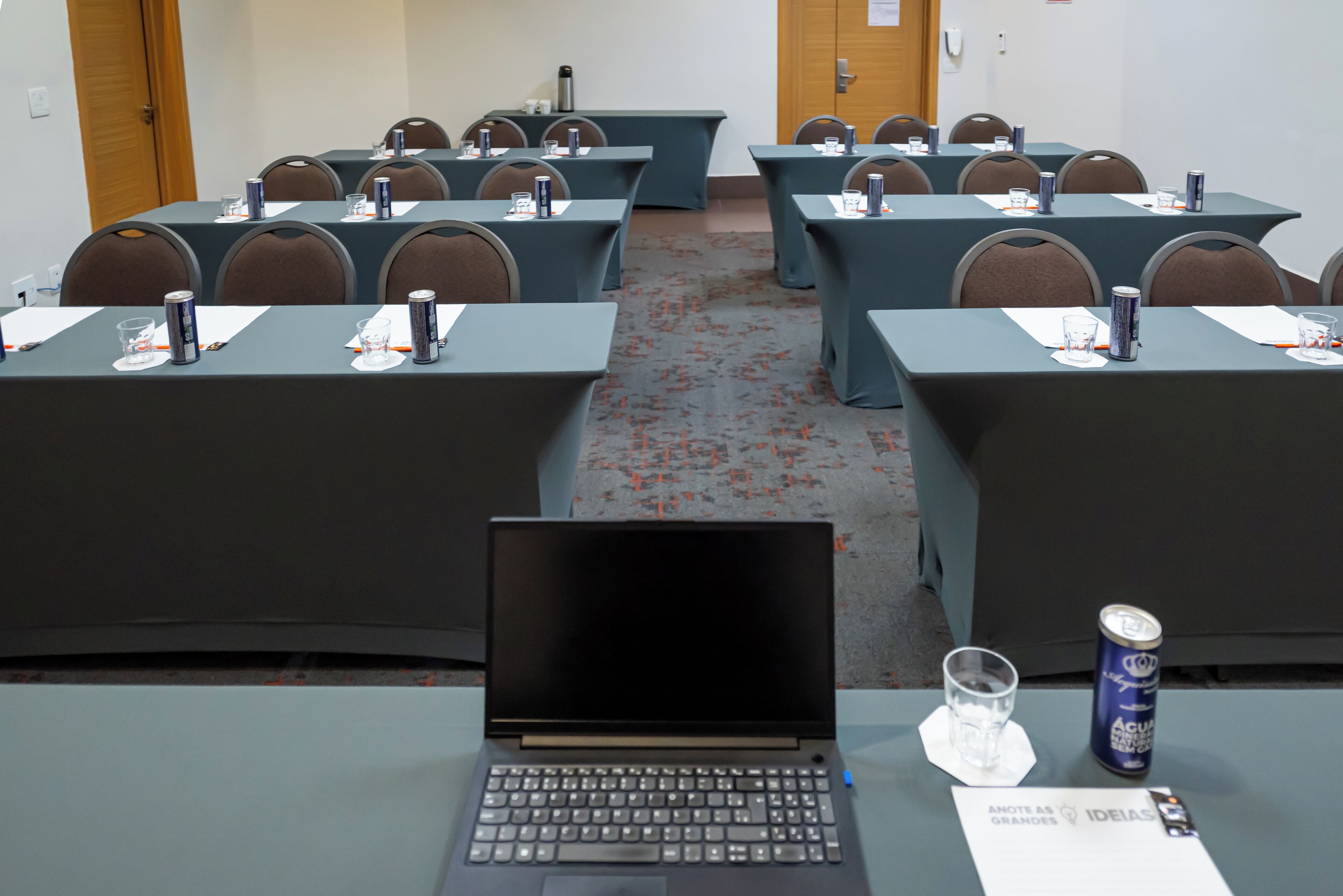 A conference room with multiple tables, each equipped with a laptop, water glass, and a can of drink.