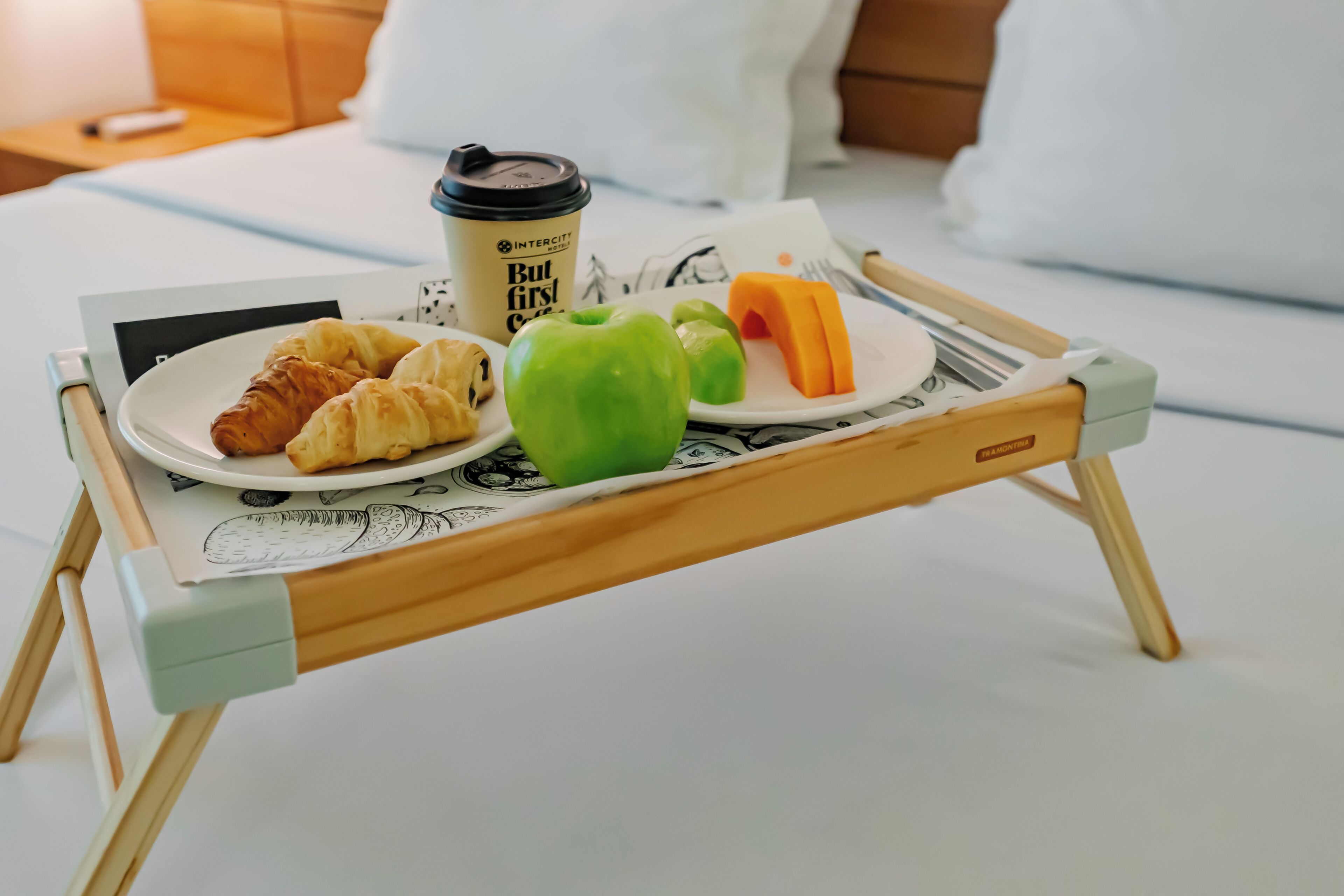 Bandeja de desayuno con cruasanes, un pimiento verde, una rodaja de naranja y una taza de café sobre una cama.