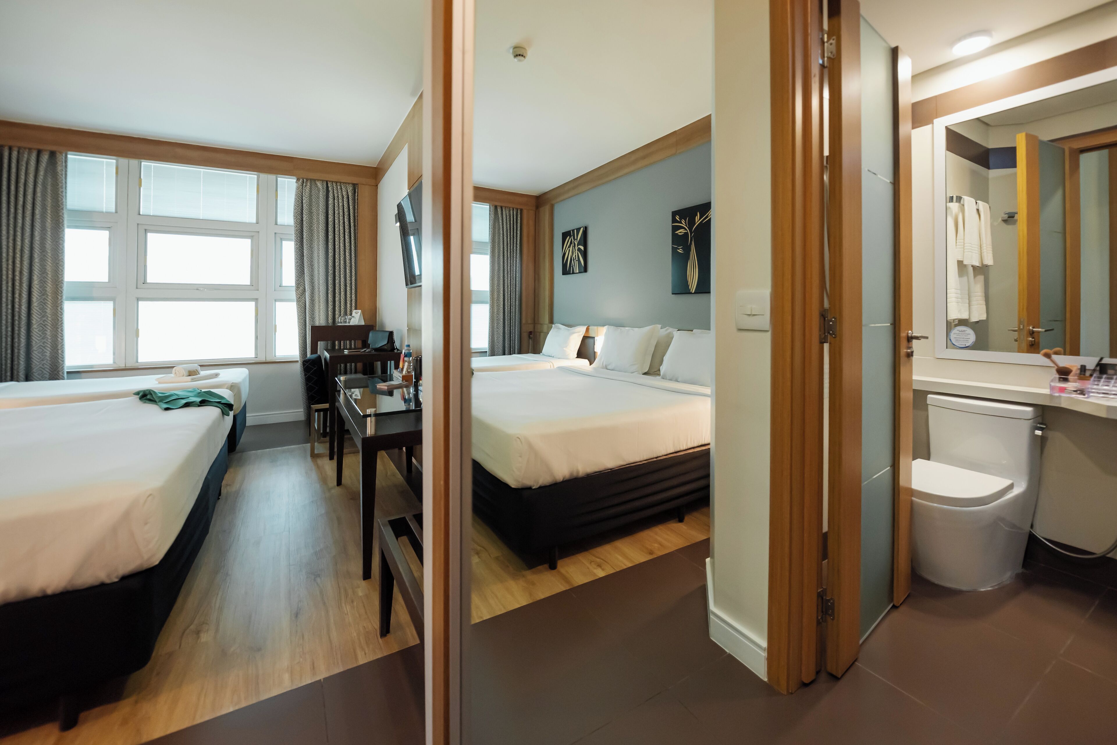 A hotel room with two beds, a desk, a TV, and a bathroom with a toilet and mirror.