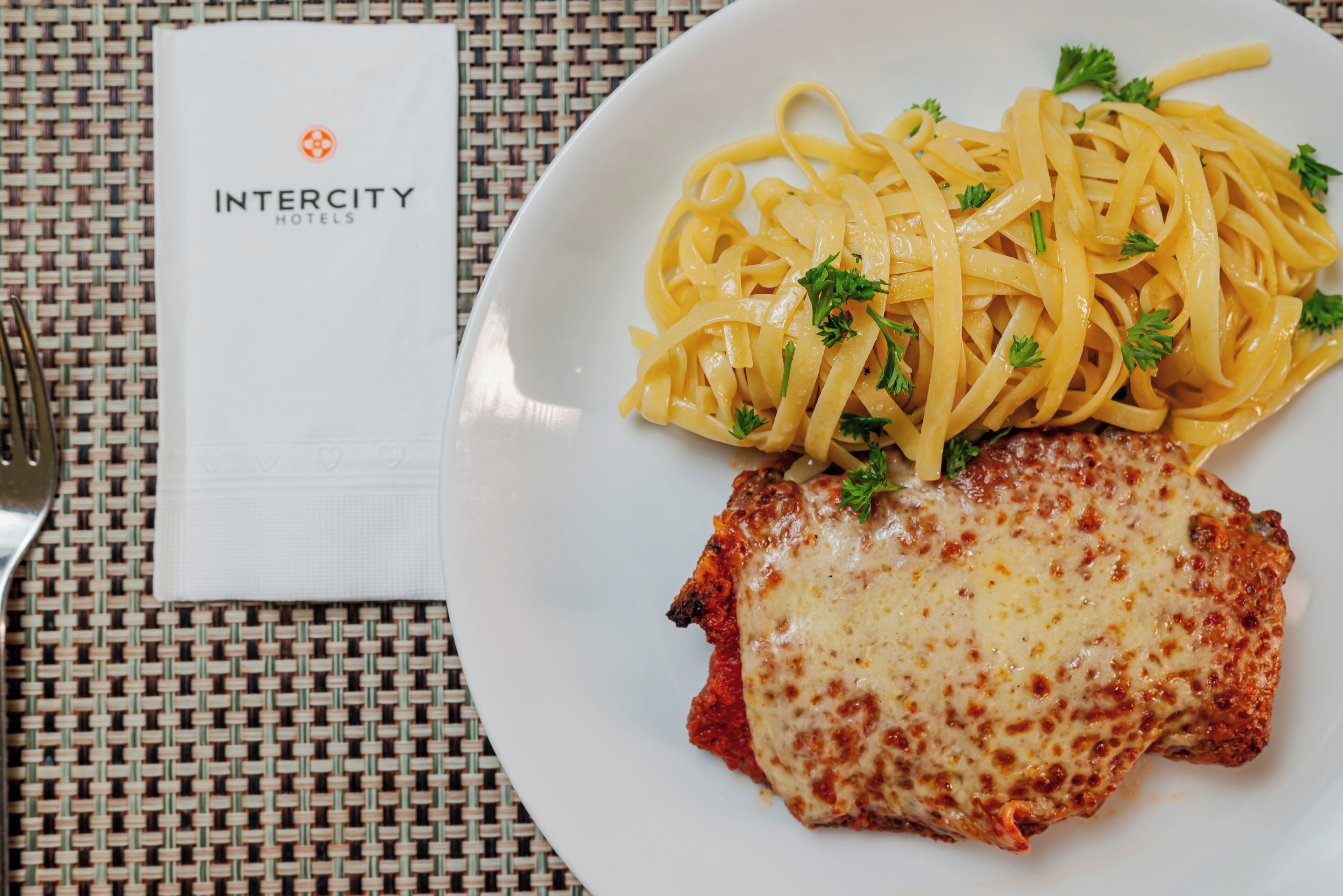 A plate of spaghetti with a meat patty and cheese, a napkin with the Intercity Hotels logo, and a fork on a woven tablecloth.