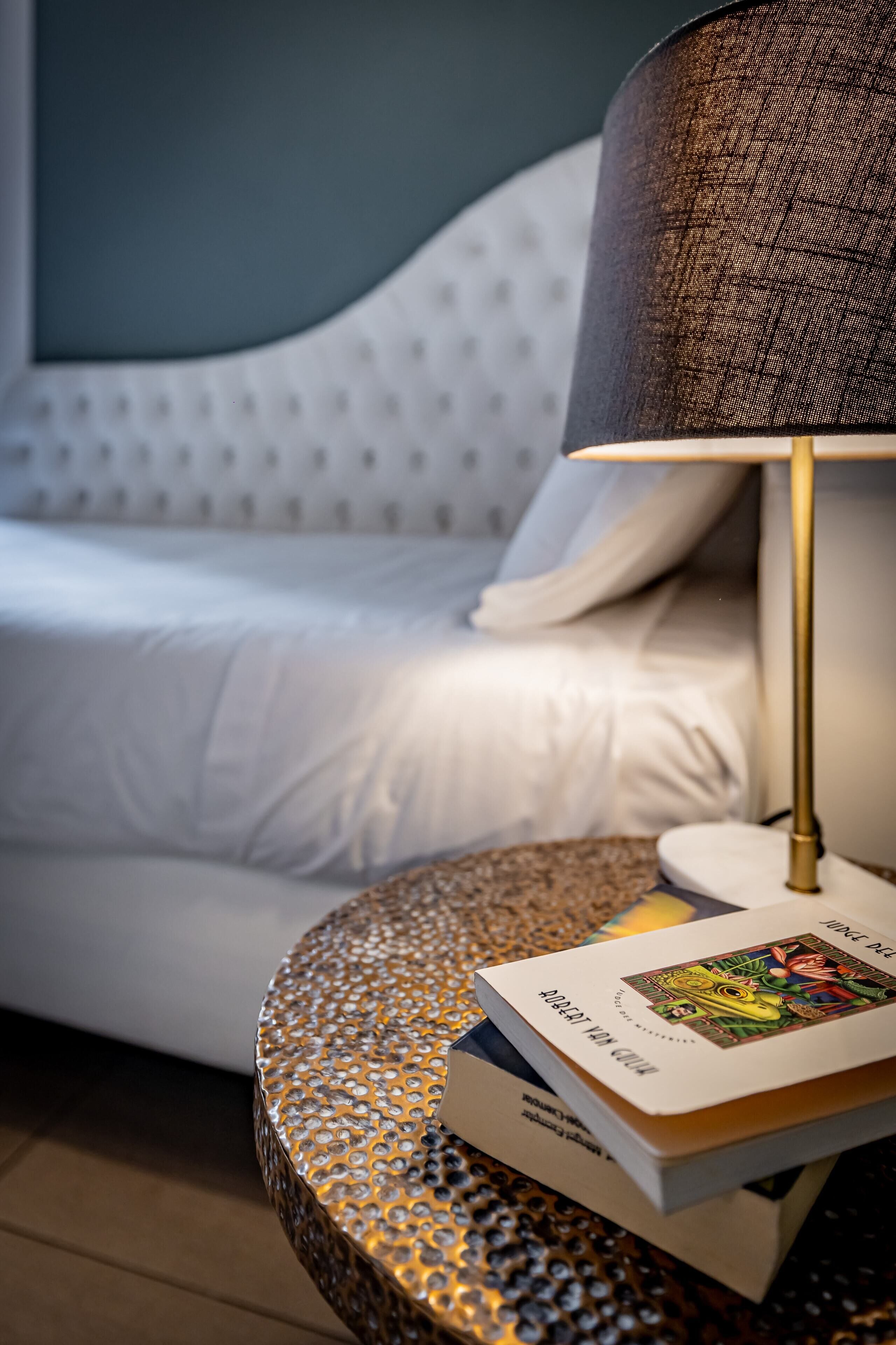 Un lit avec une literie blanche, une table d’appoint ronde à la surface texturée et une lampe avec un abat-jour foncé.