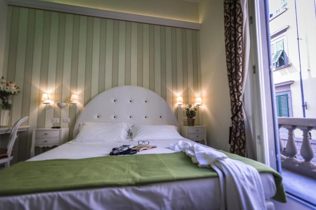 Una cama bien hecha con almohadas y una manta verde, lámparas de noche, una mesita de noche con un jarrón de flores, y vistas a un balcón con barandilla.