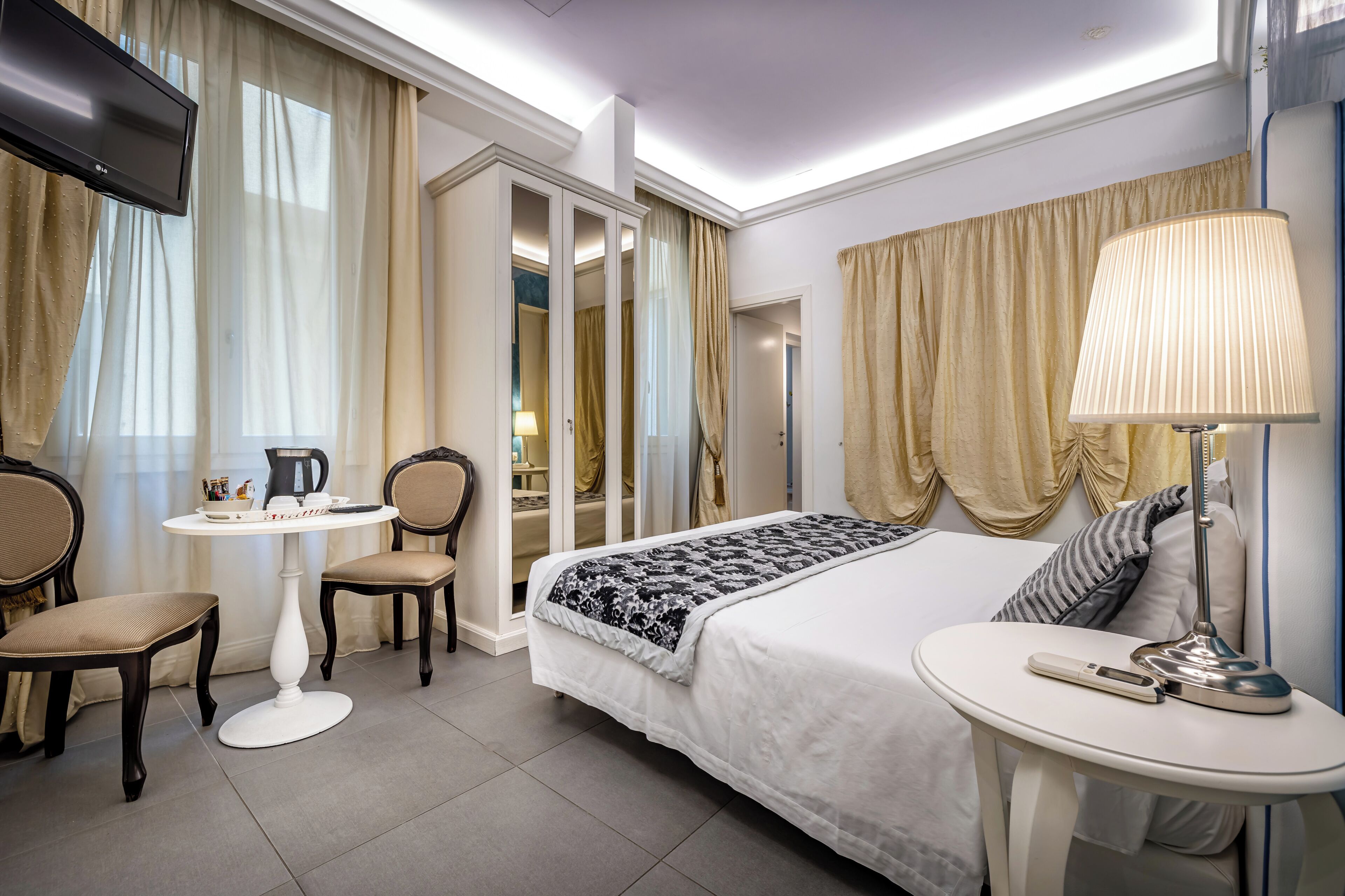 Una camera d'albergo con un letto, un comodino, una lampada, una TV e un piccolo tavolo con sedie.