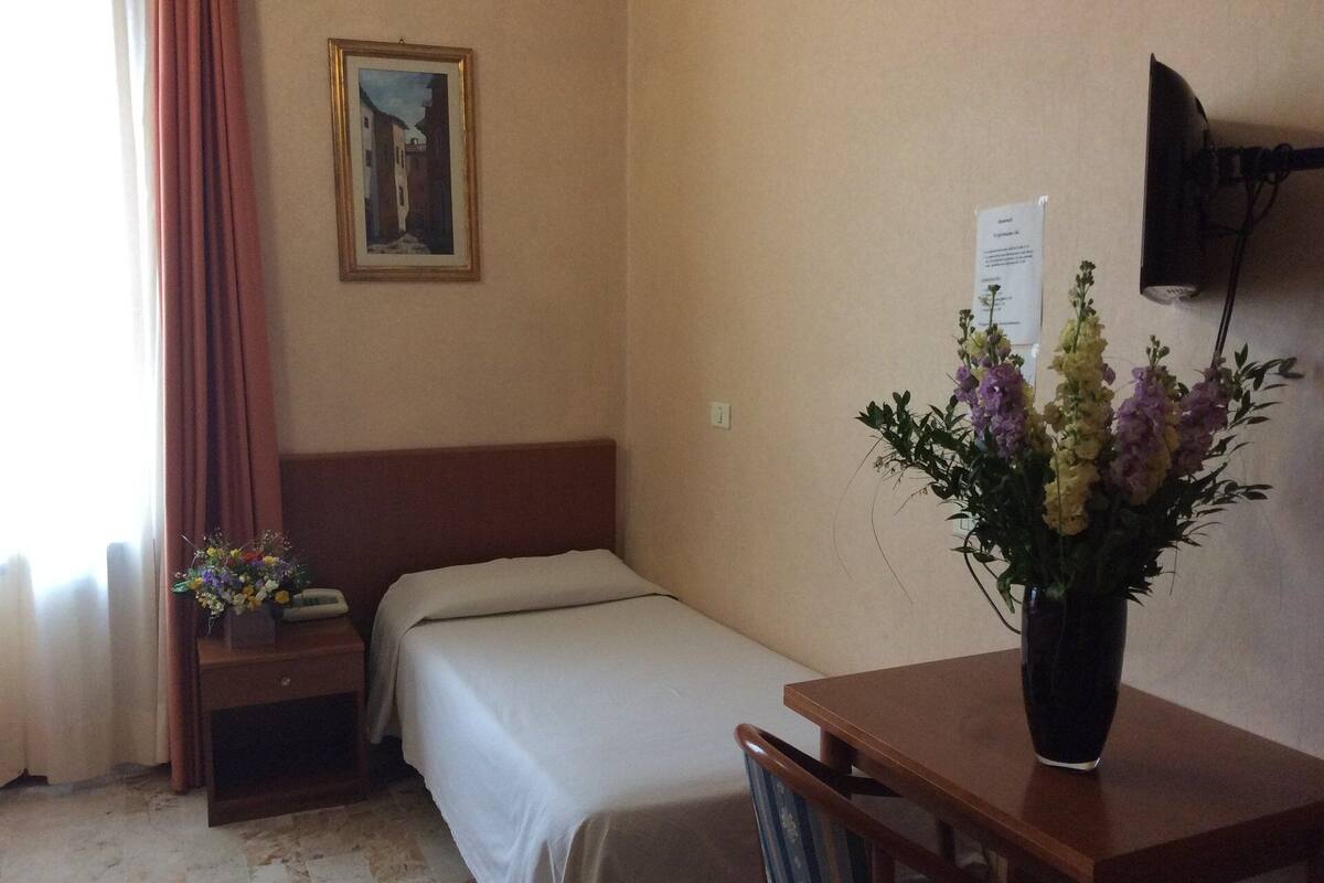 A hotel room with a single bed, a bedside table with a lamp, a small table with a vase of flowers, a painting on the wall, and a TV mounted on the wall.