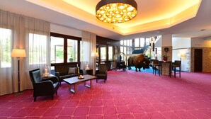 A spacious hotel lobby with a red patterned carpet, comfortable seating, a large chandelier, and a view of the outdoors. - Lobinger Hotel Parkhotel (Giengen an der Brenz)