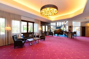Un hall d’hôtel spacieux, doté d’un tapis rouge à motifs, de sièges confortables, d’un grand lustre et offrant une vue sur l’extérieur.