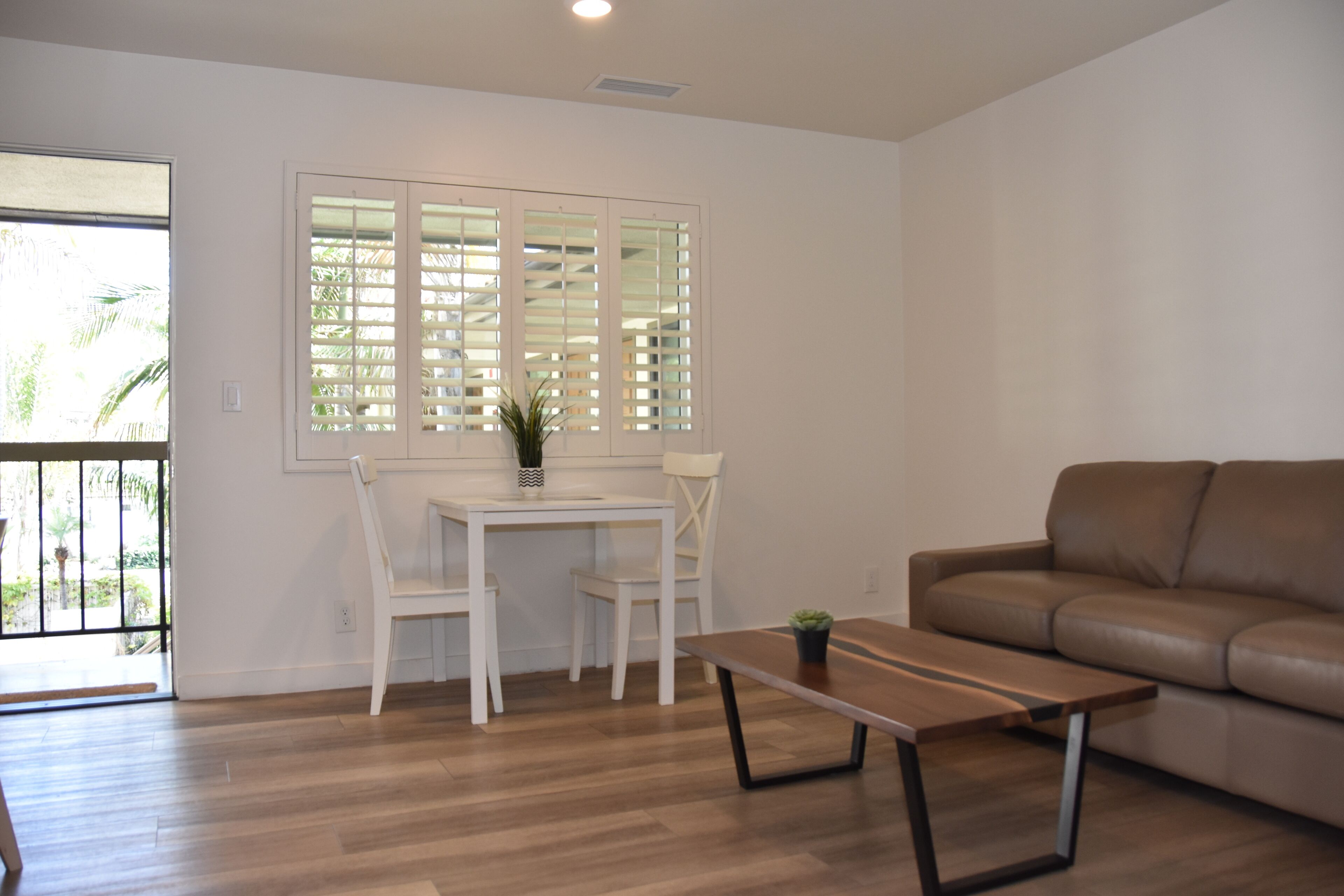 A living room with a brown leather sofa, a wooden coffee table, a white dining table with two chairs, and a window with blinds.